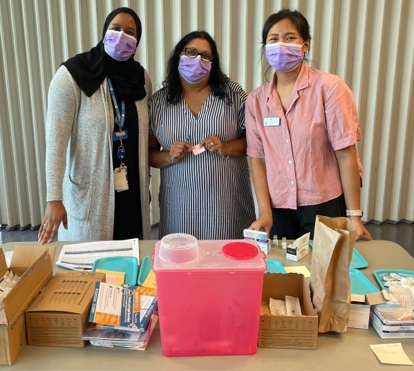 Trois femmes portant des masques violets derrière une table où se trouvent du matériel de vaccination