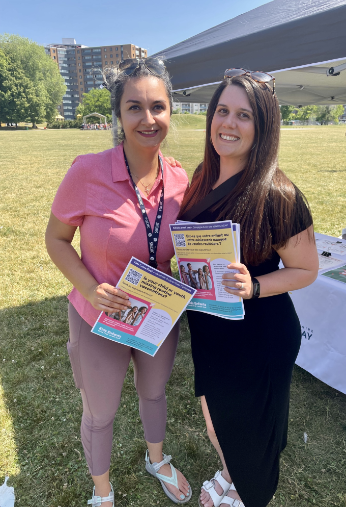 Two women holding Vaccinate & Up-to-Date posters in the park